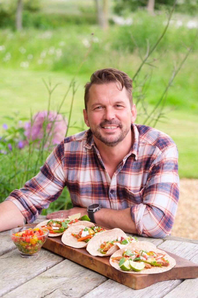 Jimmy Doherty seated at an outdoors table with a serving plate of beef fajitas and tomato salsa on the table